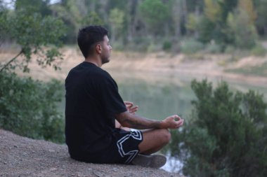 A young man sits peacefully by a tranquil lake, meditating in a calm, reflective state. The serene scene captures the harmony between body and nature clipart