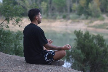 Genç bir adam sakin bir gölün kenarında huzur içinde oturur, sakin ve düşünceli bir şekilde meditasyon yapar. Huzurlu sahne, vücut ve doğa arasındaki uyumu yakalar.