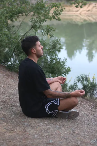 Genç bir adam sakin bir gölün kenarında huzur içinde oturur, sakin ve düşünceli bir şekilde meditasyon yapar. Huzurlu sahne, vücut ve doğa arasındaki uyumu yakalar.
