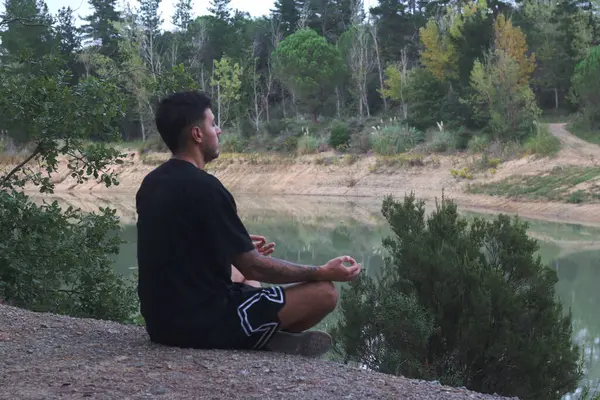 Genç bir adam sakin bir gölün kenarında huzur içinde oturur, sakin ve düşünceli bir şekilde meditasyon yapar. Huzurlu sahne, vücut ve doğa arasındaki uyumu yakalar.