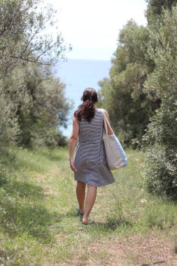 Genç bir kadının, ağaçlarla kaplı bir patikadan denize doğru yürüdüğü sakin bir sahne. Doğanın, maceranın ve sükunetin özünü yakalar.