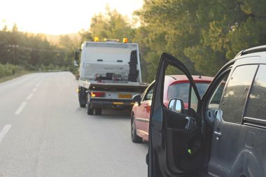 Yol kenarı sahnesinde, güvenlik için uyarı üçgenleri kurulmuş çekici bir aracın yardımıyla teknik bir sorunu olan bir araba görülüyor.