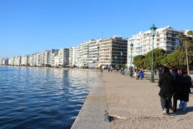 Yunanistan 'ın Selanik kentindeki deniz kenarındaki gezinti alanında, modern apartman binaları, sakin bir mavi deniz ve gezinen insanların canlı bir görüntüsü.