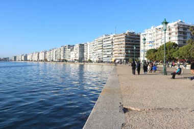 Yunanistan 'ın Selanik kentindeki deniz kenarındaki gezinti alanında, modern apartman binaları, sakin bir mavi deniz ve gezinen insanların canlı bir görüntüsü.