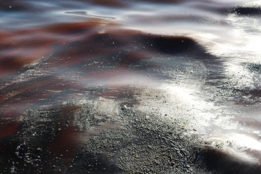Kirlenmiş bir su yüzeyinin çarpıcı bir görüntüsü turuncu, siyah ve kahverengi tonlarında dönen yağlı desenleri gösteriyor.