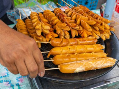 A street vendor hand is picking a bamboo sausage skewer in a process of cooking over a hot black pan.