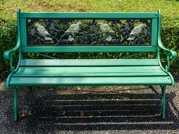A green park bench with wooden seat, iron frame, and a black backrest with four birds ornament, on a stone path at a public park.