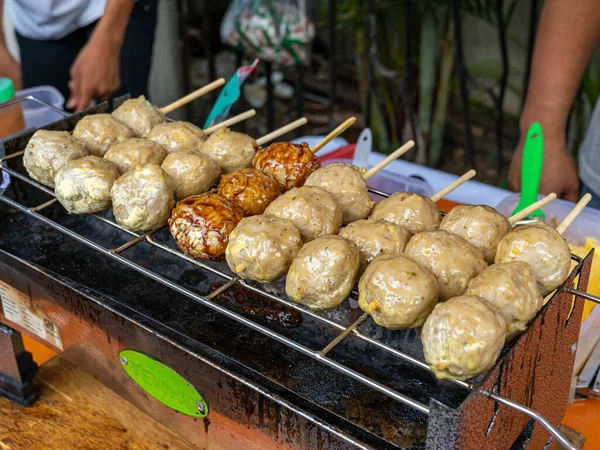 Eight meatball satays being cooked on a grilling bars, consisting of three large meatballs each.