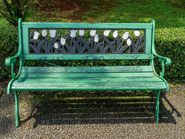 A green park bench with tulip flowers ornament, on a stone pathway during a sunny morning.