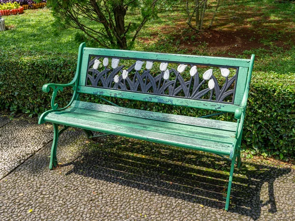 A green park bench in front of a small hedgerow and under a pine tree, with tulip flowers ornament.