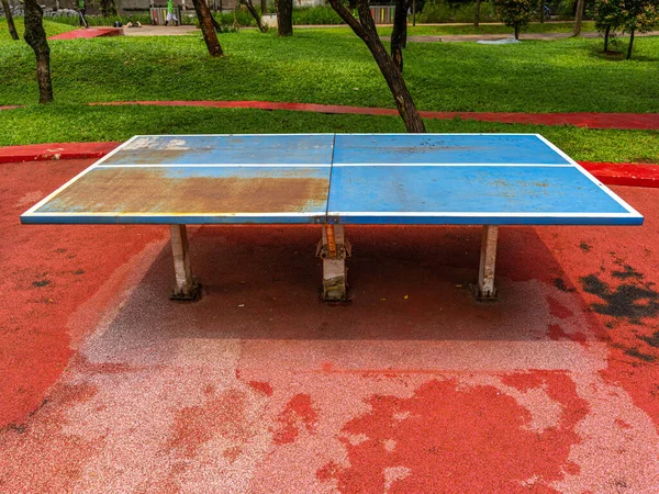 An old ping pong table with a rusty area placed on an orange cement floor at a public park.
