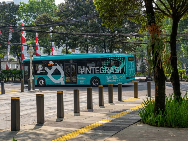 Jakarta, Indonesia - August 19, 2022: A teal blue electric city bus on service near a public park on a sunny morning.