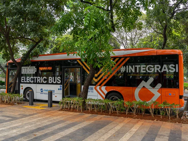 Jakarta, Indonesia - August 19, 2022: An orange and white electric city bus with open side doors waiting for passengers in front of a public park on a sunny morning.