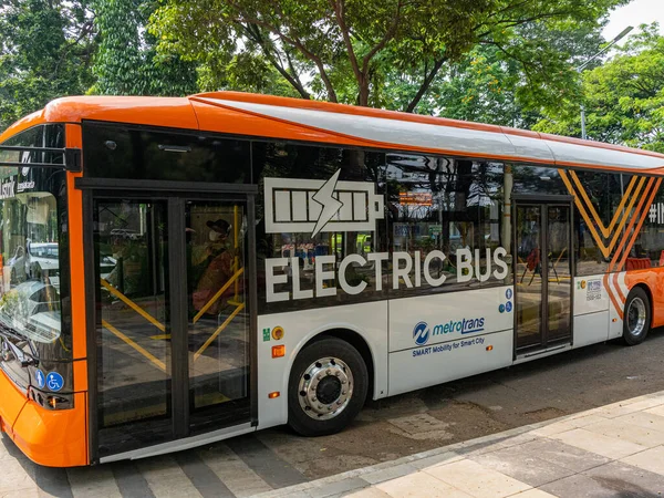 Jakarta, Indonesia - August 19, 2022: A side view of an orange and white electric city bus making a stop in front of a sidewalk shelter near a public park on a sunny morning.