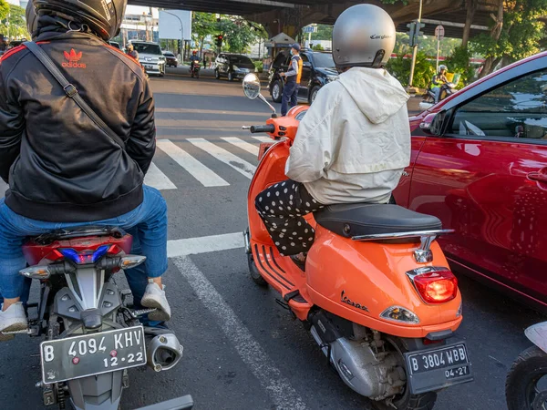 Jakarta, Indonesia - August 18, 2022: A lady riding an orange Vespa scooter at an intersection, waiting for traffic light on a sunny afternoon..