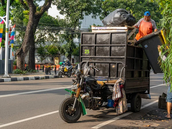 Jakarta, Indonesia - August 17, 2022: A male sanitary worker is loading garbage into his light garbage transport vehicle on a sunny morning.