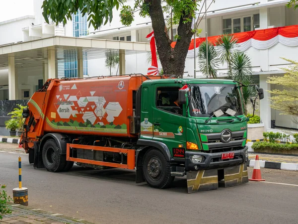 Jakarta, Indonesia - August 19, 2022: Front and side view of a garbage truck with orange and green color combination in front of a decorated building on a sunny morning.