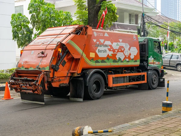 Jakarta, Indonesia - August 19, 2022: Rear and side view of a garbage truck with orange and green color combination in front of a decorated building on a sunny morning.