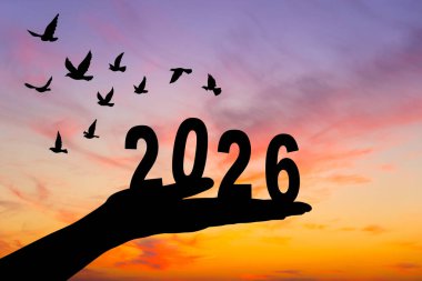 2026 rakamını kaldıran bir elin silüeti çarpıcı bir günbatımına karşı, geçişi ve yeni başlangıçları simgeliyor..
