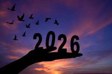 2026 rakamını kaldıran bir elin silüeti çarpıcı bir günbatımına karşı, geçişi ve yeni başlangıçları simgeliyor..