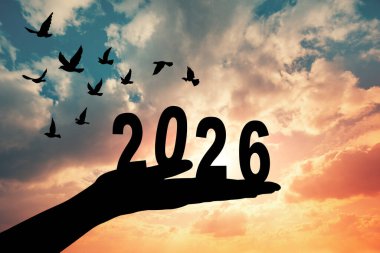 2026 rakamını kaldıran bir elin silüeti çarpıcı bir günbatımına karşı, geçişi ve yeni başlangıçları simgeliyor..