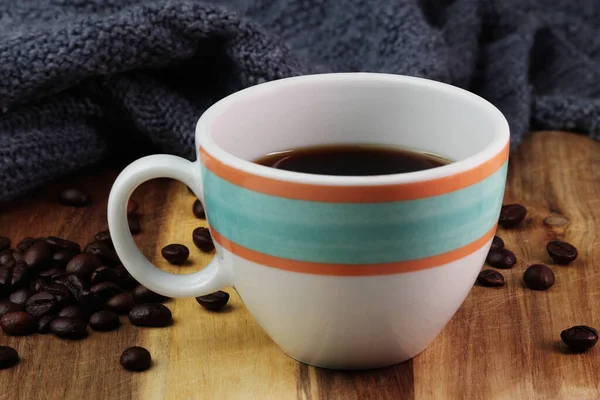 Colorful cup of black coffee is placed on a wooden surface. The beautiful cup with a handle is white with orange and aqua colored rings drawn on them. The surface has an authentic wooden texture. The shot contains a dark blue warm cozy blanket.