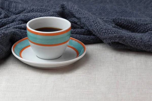 Colorful cup of black coffee is placed on the saucer. Both, the cup and the saucer are white with orange and aqua colored rings drawn on them. The surface is covered with a generic slightly creme colored blanked. The shot contains a cozy blanket.