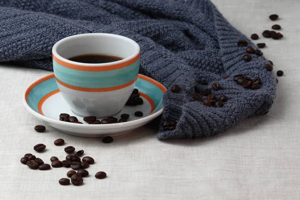 Colorful cup of black coffee is placed on the saucer. Both, the cup and the saucer are white with orange and aqua colored rings drawn on them. The surface is covered with a generic slightly creme colored blanked. The shot contains a blue blanket.