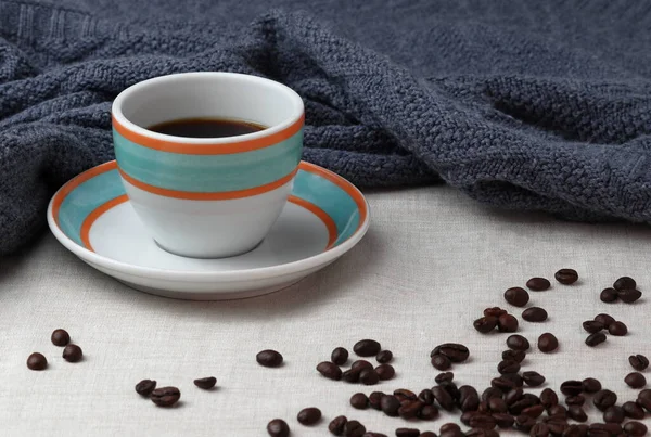 Colorful cup of black coffee is placed on the saucer. Both, the cup and the saucer are white with orange and aqua colored rings drawn on them. The surface is covered with a generic slightly creme colored blanked. The shot contains a dark blanket.
