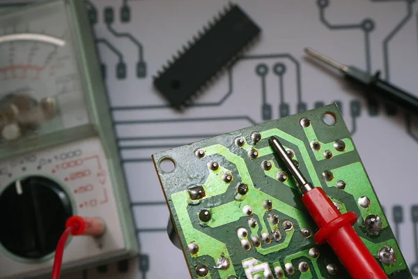 Electronic repair bench. Electronic circuit board with clurred multimeter, chip and diagram background.Focused on red probe.