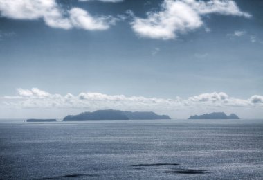 Ufuktaki Çöl Adaları 'nın silueti engin ve bulutlu bir gökyüzü tarafından çerçevelenmiştir. Deniz sakin, sakin ama biraz da gizemli bir atmosfer yaratıyor. Karanlık siluetler ve sessiz gökyüzü adaların uzak doğasını vurguluyor..