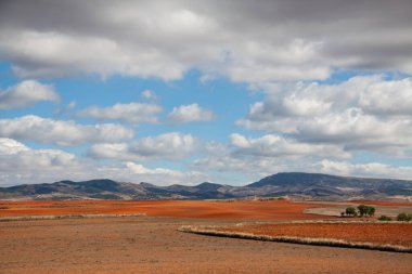 Arka planda tepeler ve bulutlu mavi gökyüzü olan kıpkırmızı topraklar..
