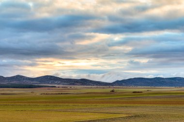 Ufukta dağlarla kaplı ekin tarlaları. Gün batımında bulutlarla kaplı görkemli mavi gökyüzü..