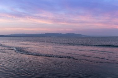 Su, gökyüzü ve kumsalda pembe karınlı dağlar.