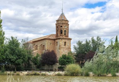 San Sebastian Kilisesi, gölün yanında bulutlarla kaplı gökyüzü olan bitki örtüsüyle çevrili. Campillo, Teruel, İspanya