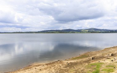 Ferbenza gölünün kıyıdan görünen gölgesi, sularına yansıyan bulutlarla kaplı yeşil tepeler. Galiçya, İspanya