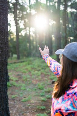 Çok renkli şapkalı ve spor ceketli Latin bir kadın ormanda güneşi işaret ediyor. dikey resim