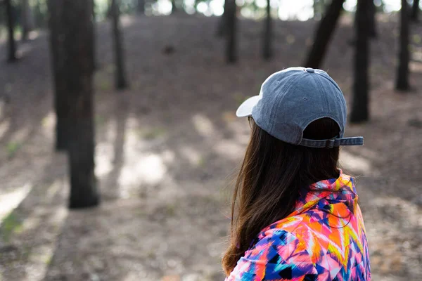 latina woman in the foreground with her back turned wearing a gray cap and a multicolored sports jacket in an autumn forest.