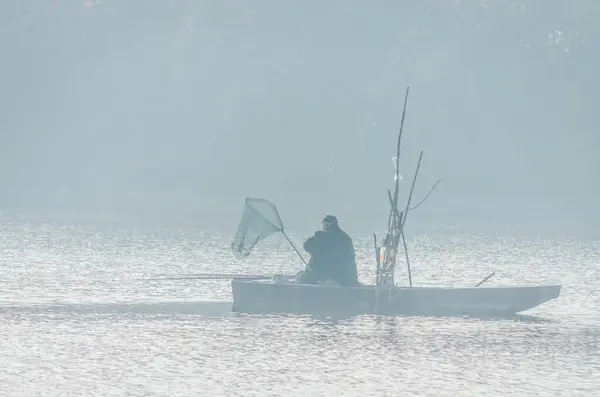 stock image A fisherman in a wooden boat on the lake, surrounded by the first morning mist and autumn sun.