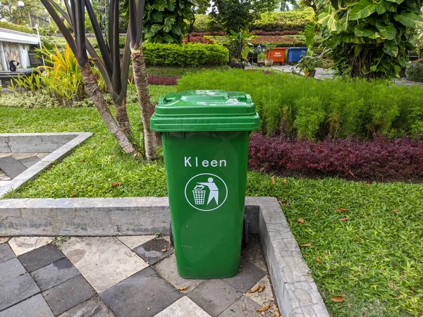 a square-shaped trash can with a green color and a garden behind it