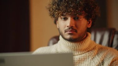 Curly bearded student studying online. Young man blinking while reading from laptop screen. Freelancer in sweater is working with computer from home. High quality 4k footage