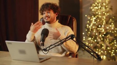 Excited streamer in sweater is gesturing during podcast session. Indoor footage of smiling influencer streaming on Christmas from home. High quality 4k footage