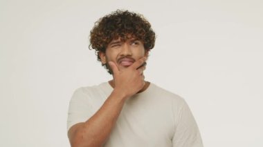 Young man makes a difficult choice. Confused male model thinking about problem. Handsome curly guy rubs his beard with bewildered face expression. High quality 4k footage