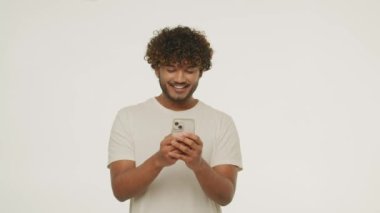 Young man in white t-shirt laughing while texting a message. Front view of a handsome guy messaging with someone with a smile. Curly male model using smartphone. Online communication. High quality 4k