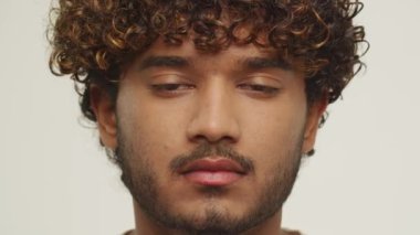 Close up view of a curly young man blinking with slight smile. Cheerful male model with dark hair is expressing positive emotion. High quality 4k footage