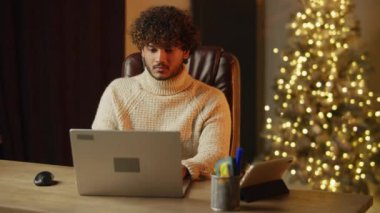 Young man in knitted sweater is working on Christmas holidays. Indoor footage of curly freelancer using laptop at his workplace with decorated Christmas tree. High quality 4k footage