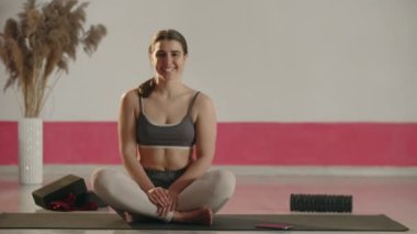 Smiling girl sitting in lotus pose and pointing with finger to something. Front view of excited young woman sitting on yoga mat after home training. High quality 4k footage