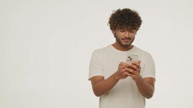 Handsome mixed man typing on smartphone and looking away with smile. Studio footage of curly guy in t-shirt messaging and laughing. Excited male model texting a message with positive emotions. High