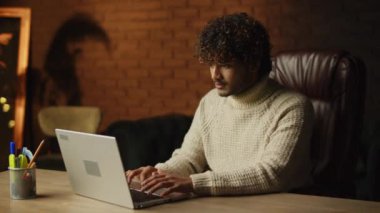 Focused freelancer typing on laptop keyboard. Tired young man in sweater working from home. High quality 4k footage