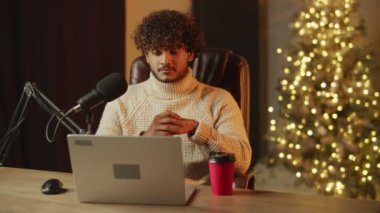 Curly blogger is nodding while listening podcast and drinking coffee. Online influencer is doing live stream on Christmas. High quality 4k footage